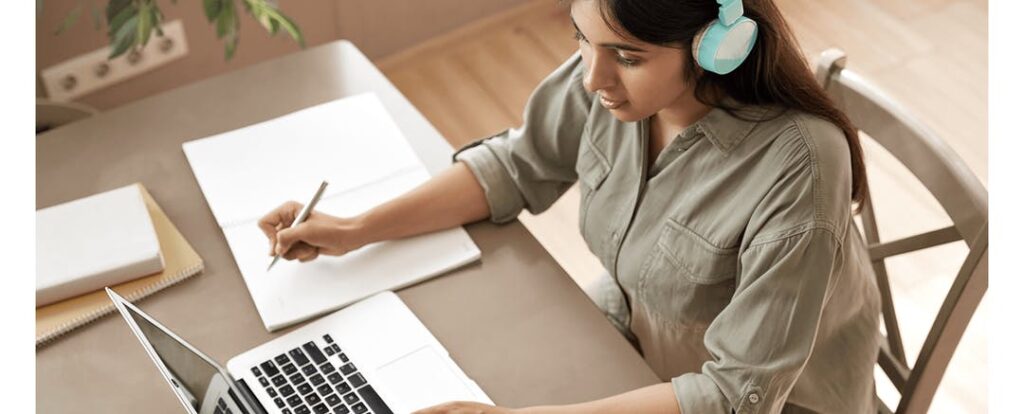 Diligent online student wearing blue headphones and writing in a notebook while viewing online content on a laptop
